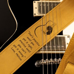 K&ouml;nnte beinhalten: Ein hellbrauner Gitarrengurt aus Leder mit einer schwarzen Gitarre. Der Gurt hat eine Gitarrengrafik und das Zitat: &bdquo;&Uuml;be nicht, bis du es richtig kannst, &uuml;be, bis du es nicht mehr falsch machen kannst.&ldquo; Der Gurt liegt auf einer schwarzen Gitarre.