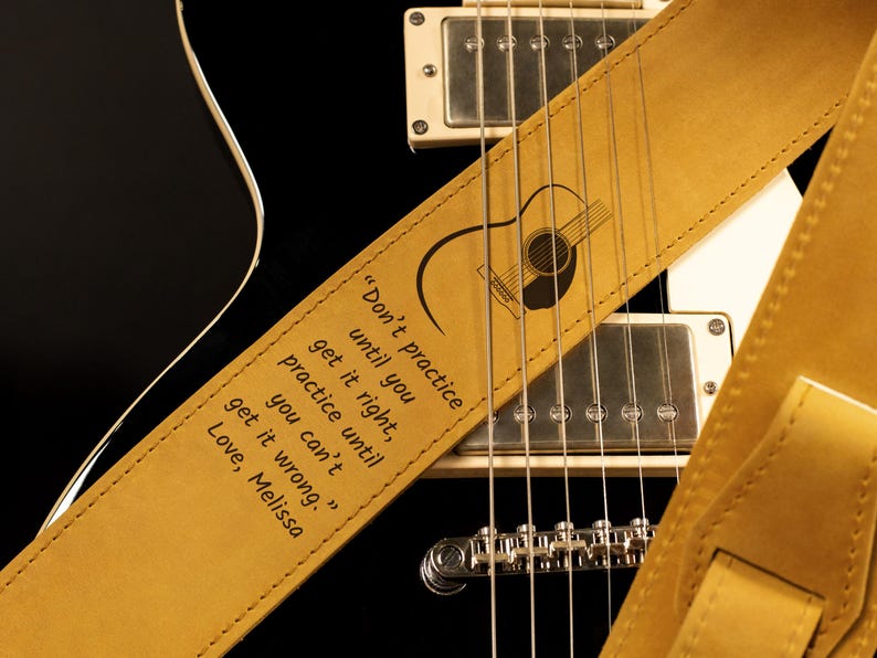 K&ouml;nnte beinhalten: Ein hellbrauner Gitarrengurt aus Leder mit einer schwarzen Gitarre. Der Gurt hat eine Gitarrengrafik und das Zitat: &bdquo;&Uuml;be nicht, bis du es richtig kannst, &uuml;be, bis du es nicht mehr falsch machen kannst.&ldquo; Der Gurt liegt auf einer schwarzen Gitarre.