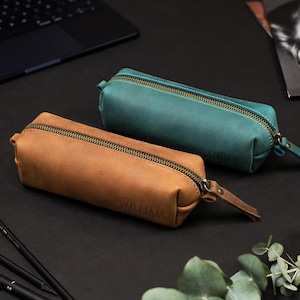 May include: Two leather pencil cases, one in a warm brown and the other in teal, with brass zippers and matching pull tabs. The brown case is embossed with the name "WILLIAM". The cases are on a black surface, with pens and a laptop visible in the background.