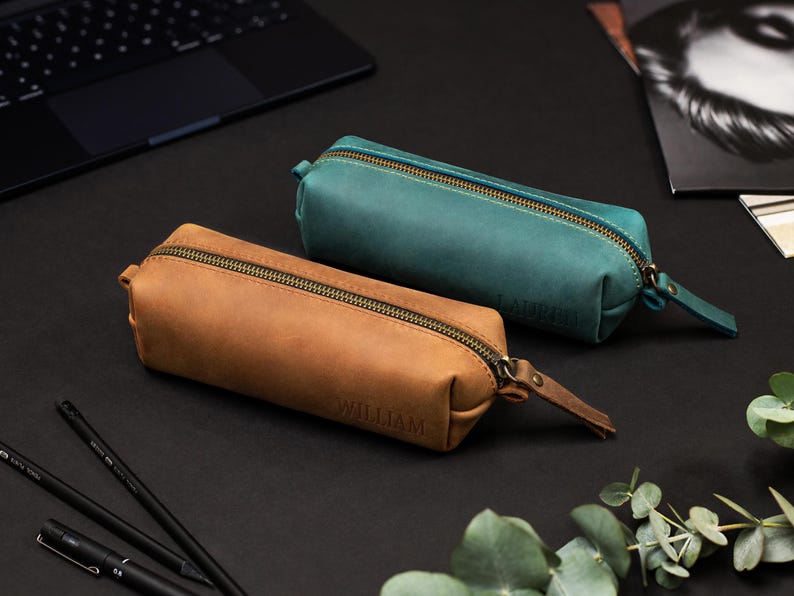 May include: Two leather pencil cases, one in a warm brown and the other in teal, with brass zippers and matching pull tabs. The brown case is embossed with the name "WILLIAM". The cases are on a black surface, with pens and a laptop visible in the background.