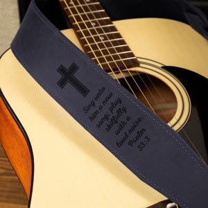 K&ouml;nnte beinhalten: Akustikgitarre mit einem marineblauen Gitarrengurt. Der Gurt zeigt ein schwarzes Kreuz und den Text "Singet ihm ein neues Lied; spielt ihm herrlich auf mit Jubel. Psalm 33,3."