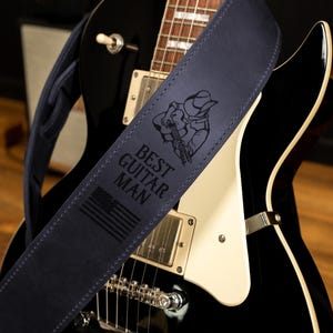 May include: A navy blue guitar strap with the text "BEST GUITAR MAN" and an illustration of a cowboy playing a guitar. The strap also features an American flag design. The strap is draped over a black electric guitar.