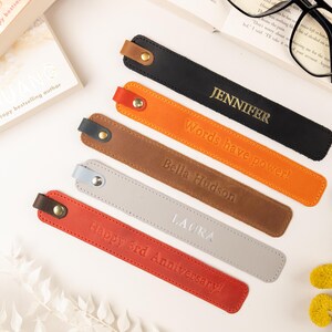 May include: Leather bookmarks in black, orange, brown, gray, and red, each with a different name or message embossed in gold or white. The bookmarks are arranged near a book and glasses.