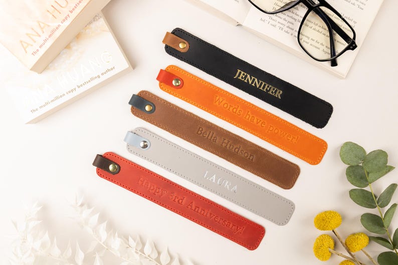 May include: Leather bookmarks in black, orange, brown, gray, and red, each with a different name or message embossed in gold or white. The bookmarks are arranged near a book and glasses.
