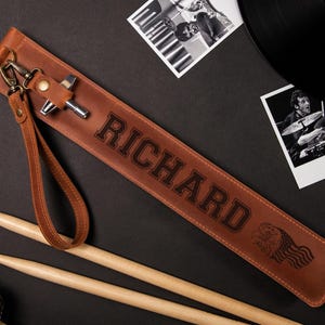 May include: A brown leather drumstick case with the name "RICHARD" embossed in large letters. The case has a wrist strap and a metal clasp. Two drumsticks and a microphone are also visible.