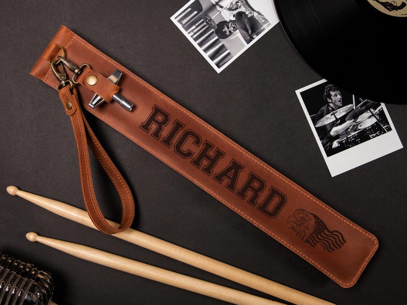May include: A brown leather drumstick case with the name "RICHARD" embossed in large letters. The case has a wrist strap and a metal clasp. Two drumsticks and a microphone are also visible.