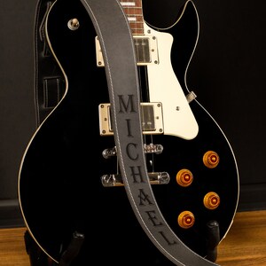 May include: A black electric guitar with a white pickguard and gold hardware. A gray leather guitar strap with the name "MICHAEL" is draped over the guitar. The guitar is on a black stand.