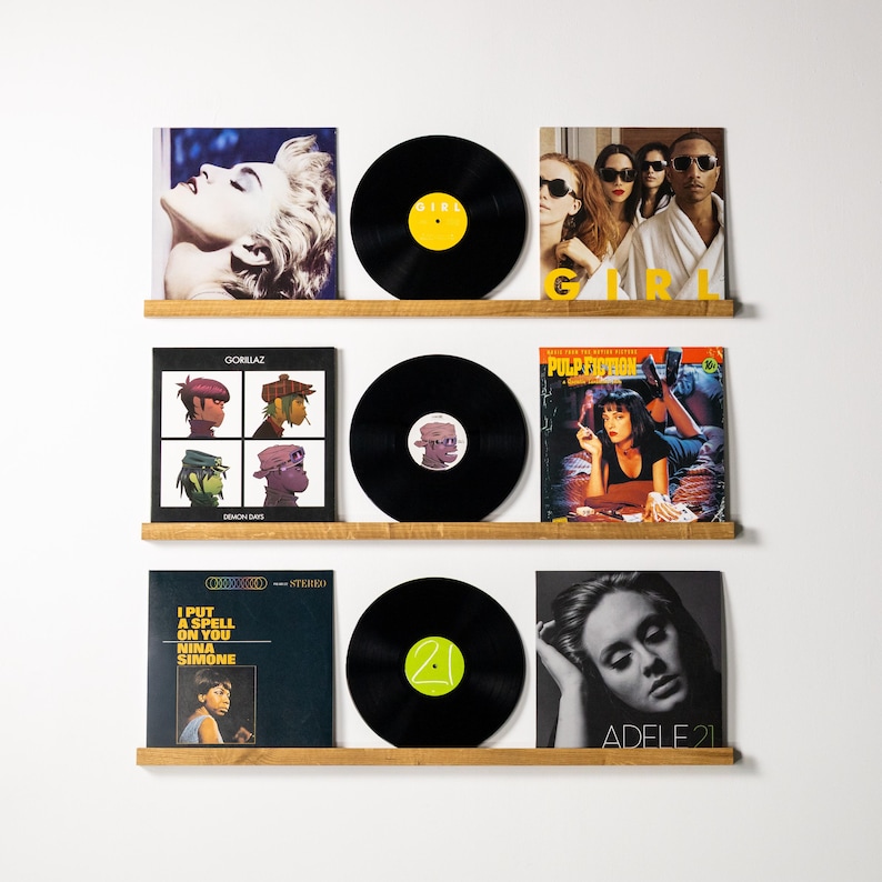 May include: A collection of vinyl records displayed on wooden shelves against a white wall. The album covers feature diverse artwork and artist portraits. The records are black with colorful labels. The albums include Madonna, Gorillaz, Pulp Fiction, Nina Simone, and Adele.