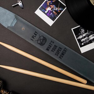 May include: A gray leather drumstick case with the text "I PLAY WHAT'S YOUR SUPER POWER?" and a drum symbol. Two wooden drumsticks are next to the case. The background includes a vinyl record, photos, and a drum key.