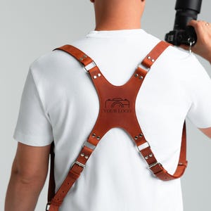 May include: A brown leather camera harness with silver metal buckles and rivets. The harness features a central panel with a camera logo and the text "YOUR LOGO". Designed to support a camera.