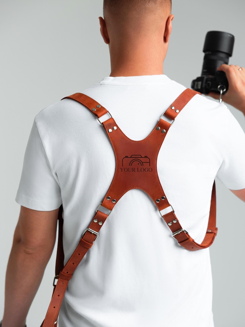 May include: A brown leather camera harness with silver metal buckles and rivets. The harness features a central panel with a camera logo and the text "YOUR LOGO". Designed to support a camera.