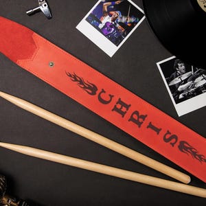 May include: A red leather drumstick case with the name "CHRIS" in black lettering and flame graphics. Two wooden drumsticks are next to the case. The background includes photos and a vinyl record.