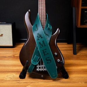 May include: A dark green leather guitar strap with the name "JAMES" printed in black. The strap is draped over a brown bass guitar. The guitar is on a wooden surface, with a black background.