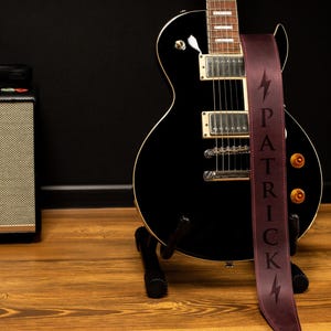 K&ouml;nnte beinhalten: Eine schwarze E-Gitarre mit einem weinroten Gurt, auf dem der Name "PATRICK" vertikal und ein Blitz abgebildet sind. Die Gitarre steht auf einem St&auml;nder, im Hintergrund sind ein Verst&auml;rker und Kopfh&ouml;rer zu sehen.
