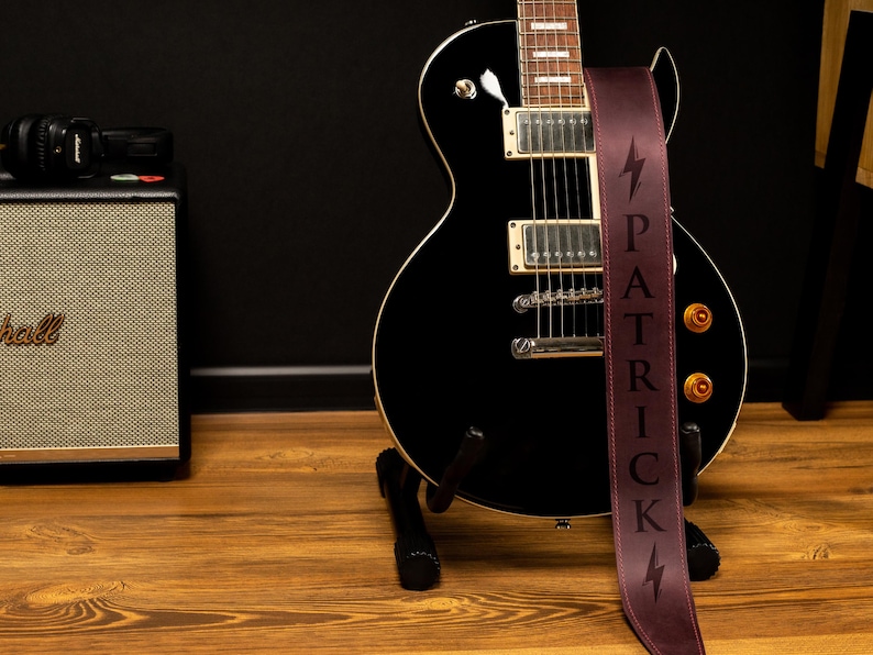 K&ouml;nnte beinhalten: Eine schwarze E-Gitarre mit einem weinroten Gurt, auf dem der Name "PATRICK" vertikal und ein Blitz abgebildet sind. Die Gitarre steht auf einem St&auml;nder, im Hintergrund sind ein Verst&auml;rker und Kopfh&ouml;rer zu sehen.