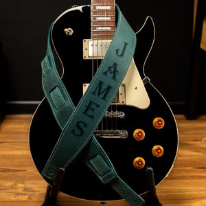 Puede incluir: Una guitarra el&eacute;ctrica negra con herrajes dorados y una correa de guitarra de cuero verde azulado. La correa est&aacute; personalizada con el nombre "JAMES" en may&uacute;sculas. La guitarra est&aacute; sobre un soporte de madera.