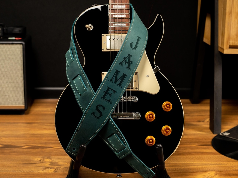 Puede incluir: Una guitarra el&eacute;ctrica negra con herrajes dorados y una correa de guitarra de cuero verde azulado. La correa est&aacute; personalizada con el nombre "JAMES" en may&uacute;sculas. La guitarra est&aacute; sobre un soporte de madera.
