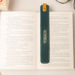 May include: A dark green leather bookmark with the name "SOPHIA" in gold lettering. The bookmark is inserted into an open book. The bookmark has a tan leather tab at the top. A pair of glasses is on the table.