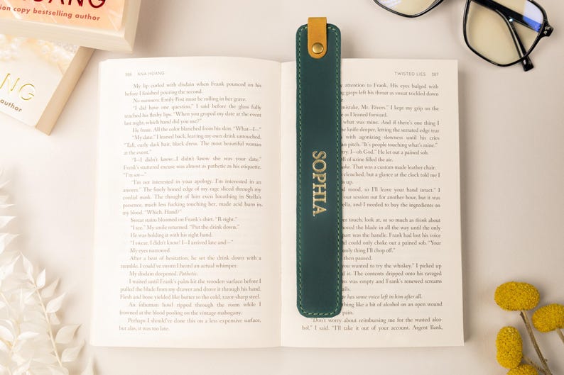 May include: A dark green leather bookmark with the name "SOPHIA" in gold lettering. The bookmark is inserted into an open book. The bookmark has a tan leather tab at the top. A pair of glasses is on the table.