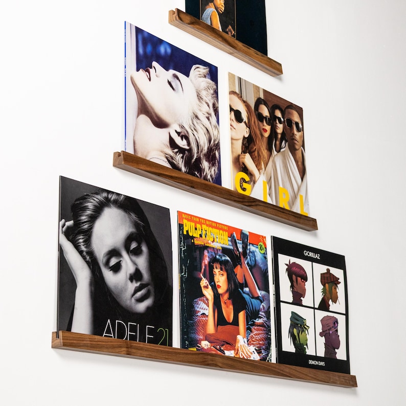 May include: Wooden floating shelves displaying vinyl record album covers. The albums feature various artists and album art, including Adele's "21", Pulp Fiction, and Gorillaz's "Demon Days". The shelves are mounted on a white wall.
