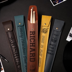 Puede incluir: Cinco estuches de cuero para baquetas de batería con diferentes diseños y texto personalizado. Los estuches son marrones, verdes, amarillos y grises. El texto en los estuches incluye "Richard", "Born to Play Drums", "Merry Christmas Dad! Rock On", "Joshua" y "I Play What's Your Super Power?"