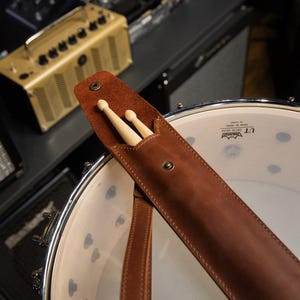 May include: A brown leather drumstick holder with two wooden drumsticks inside. The holder is attached to a drum. The background includes amplifiers and other musical equipment.