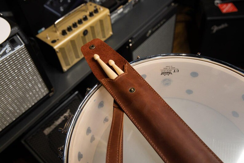 May include: A brown leather drumstick holder with two wooden drumsticks inside. The holder is attached to a drum. The background includes amplifiers and other musical equipment.