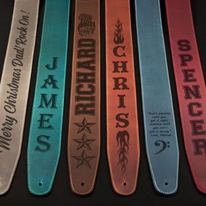 Puede incluir: Una colección de correas de guitarra personalizadas en varios colores, incluyendo gris, turquesa, marrón, naranja, azul y rojo. Cada correa presenta texto grabado, como nombres y frases. Las correas son de cuero.