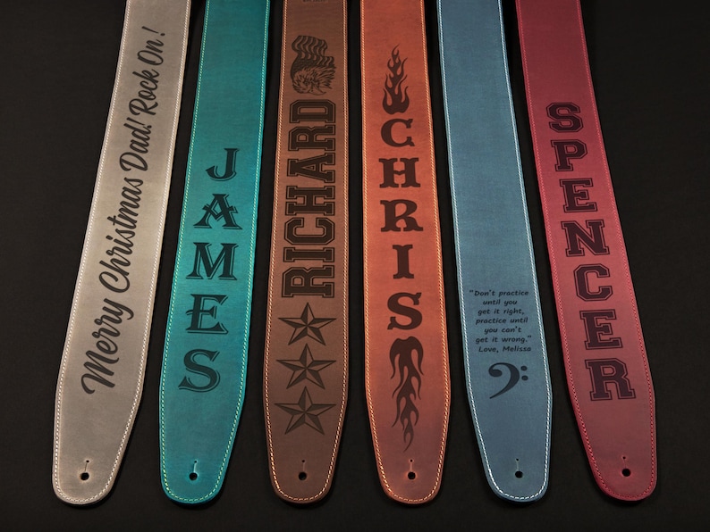 May include: A collection of personalized guitar straps in various colors, including gray, teal, brown, orange, blue, and red. Each strap features engraved text, such as names, phrases, and a musical quote. The straps are made of leather.