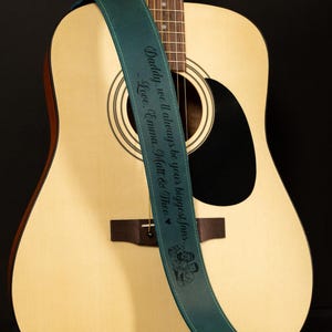 K&ouml;nnte beinhalten: Akustikgitarre mit einem tealfarbenen Gitarrengurt. Der Gurt tr&auml;gt den Text "Daddy, we'll always be your biggest fans. Love, Emma, Matt & Theo" in Schwarz. Die Gitarre ist hellbraun mit einem schwarzen Schallloch.