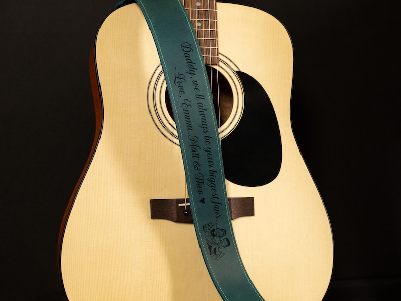 K&ouml;nnte beinhalten: Akustikgitarre mit einem tealfarbenen Gitarrengurt. Der Gurt tr&auml;gt den Text "Daddy, we'll always be your biggest fans. Love, Emma, Matt & Theo" in Schwarz. Die Gitarre ist hellbraun mit einem schwarzen Schallloch.