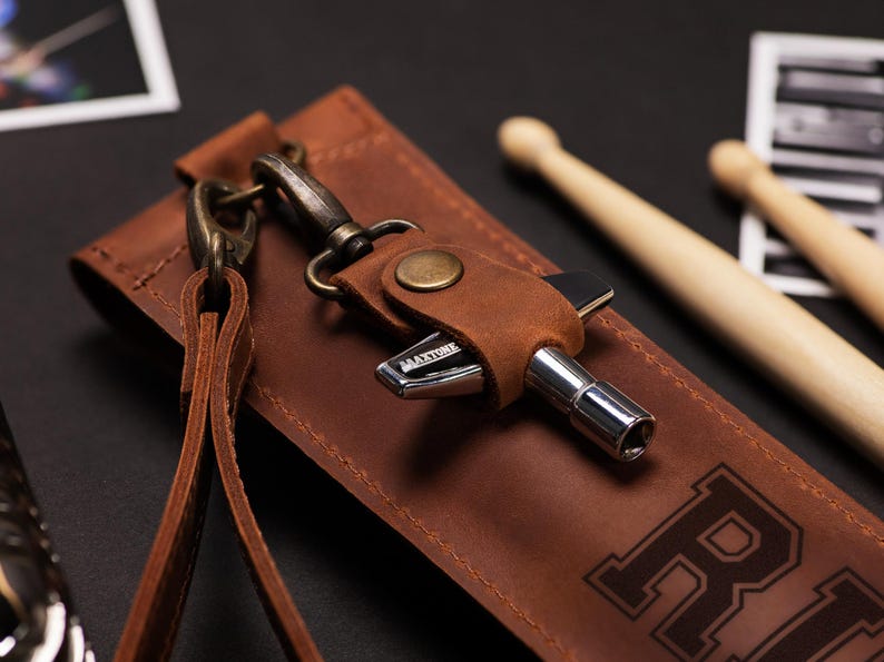 May include: A brown leather drum key holder with a metal drum key and a strap. The holder has a snap closure and a metal clasp. The word "MAXTONE" is visible on the drum key. Two wooden drumsticks are in the background.