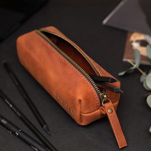 May include: A brown leather pencil case with a zipper, containing pencils. The word "RICHARD" is embossed on the side. The pencil case is open, revealing the interior. The zipper and pull tab are a contrasting color.