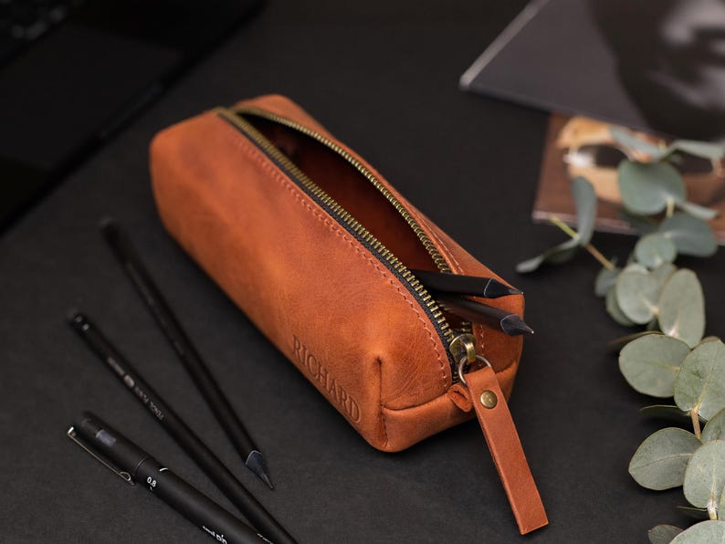 May include: A brown leather pencil case with a zipper, containing pencils. The word "RICHARD" is embossed on the side. The pencil case is open, revealing the interior. The zipper and pull tab are a contrasting color.