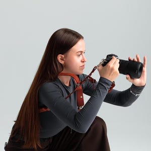 May include: A brown leather camera harness worn over a dark grey long-sleeved top. The harness features multiple straps and metal hardware. A black camera with a large lens is being held. The photographer is wearing brown trousers.