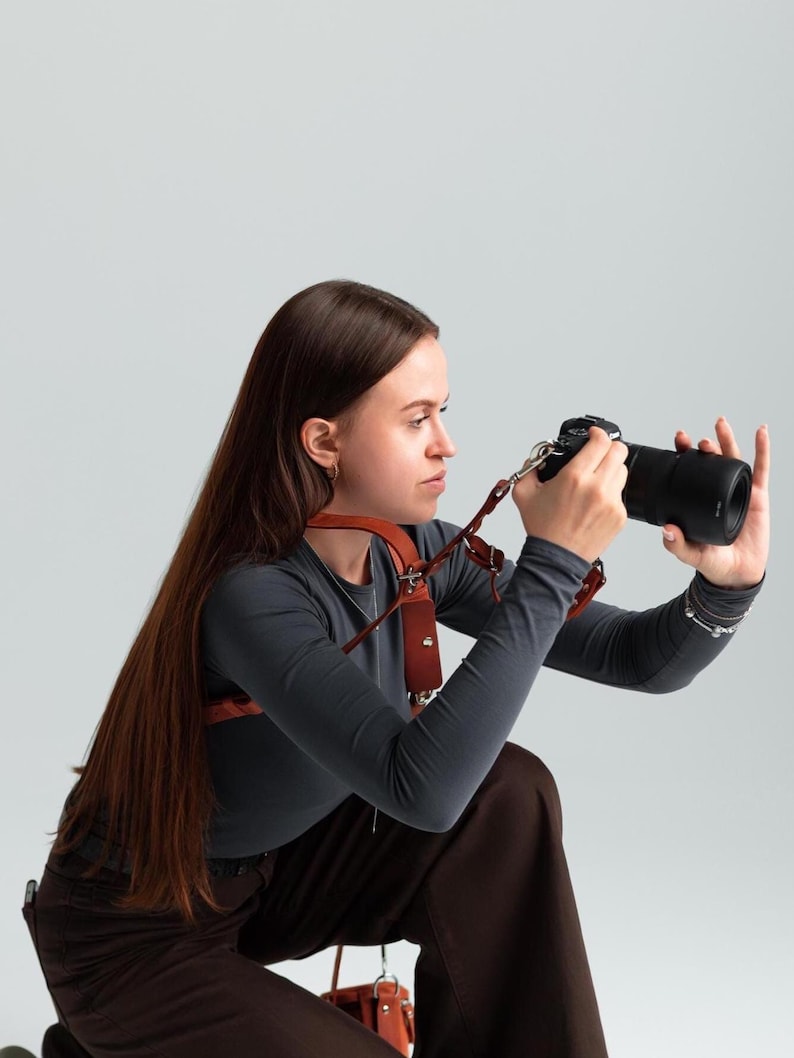 May include: A brown leather camera harness worn over a dark grey long-sleeved top. The harness features multiple straps and metal hardware. A black camera with a large lens is being held. The photographer is wearing brown trousers.