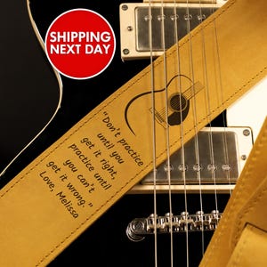 May include: A tan leather guitar strap with a guitar silhouette and the quote "Don't practice until you get it right, practice until you can't get it wrong." The strap is on a black electric guitar.