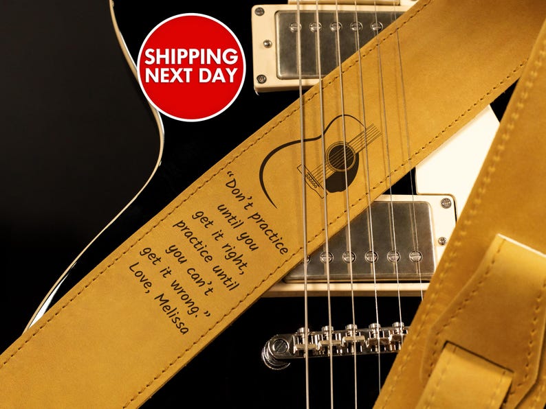 May include: A tan leather guitar strap with a guitar silhouette and the quote "Don't practice until you get it right, practice until you can't get it wrong." The strap is on a black electric guitar.