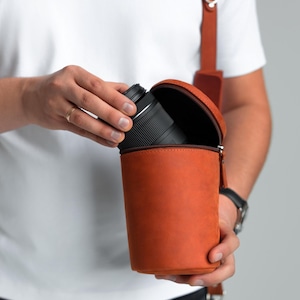 May include: A cylindrical, orange leather camera lens case with a zip closure. The case is held open, revealing a black camera lens. A brown leather strap is attached for carrying.