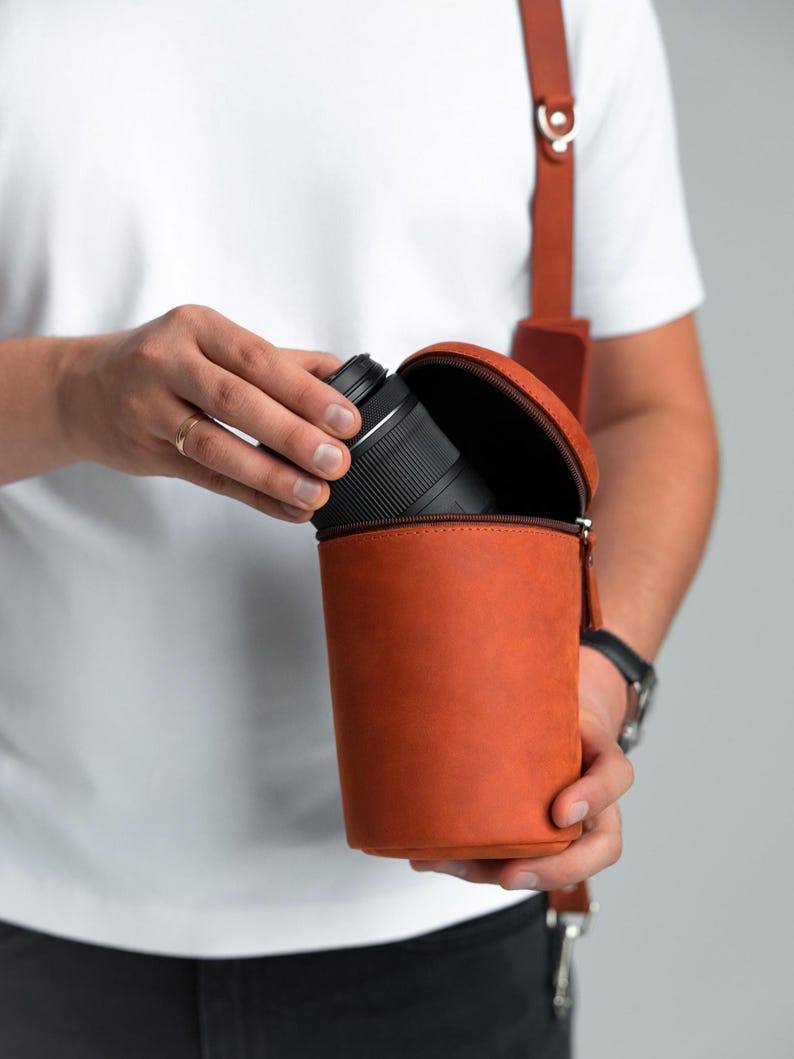 May include: A cylindrical, orange leather camera lens case with a zip closure. The case is held open, revealing a black camera lens. A brown leather strap is attached for carrying.
