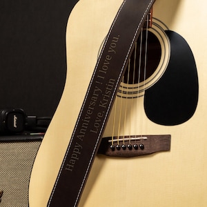 Puede incluir: Guitarra ac&uacute;stica con una correa de cuero marr&oacute;n. La correa tiene costuras blancas y texto grabado que dice "Happy Anniversary! I love you. Love, Kristin." La guitarra es de color claro con una boca negra.