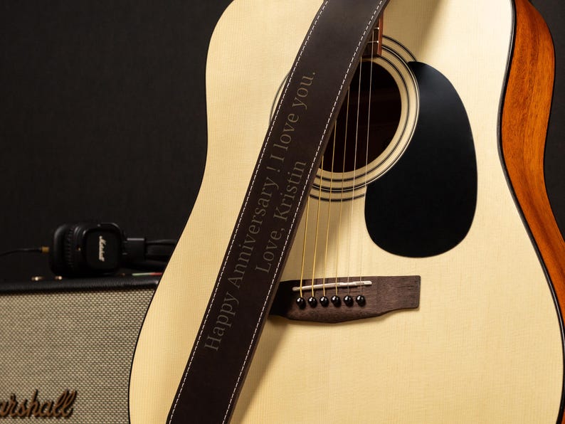 Puede incluir: Guitarra ac&uacute;stica con una correa de cuero marr&oacute;n. La correa tiene costuras blancas y texto grabado que dice "Happy Anniversary! I love you. Love, Kristin." La guitarra es de color claro con una boca negra.