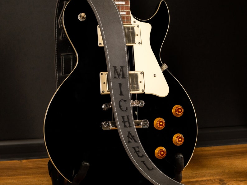 K&ouml;nnte beinhalten: Eine schwarze E-Gitarre mit wei&szlig;em Schlagbrett und goldfarbener Hardware. Ein grauer Ledergitarrengurt mit dem Namen "MICHAEL" ist &uuml;ber die Gitarre gelegt. Die Gitarre steht auf einem schwarzen St&auml;nder.