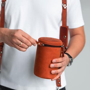 May include: A cylindrical, brown leather bag with a black zip is held by a person. The bag is worn with brown leather braces. The person is wearing a white t-shirt and a watch with a black strap.