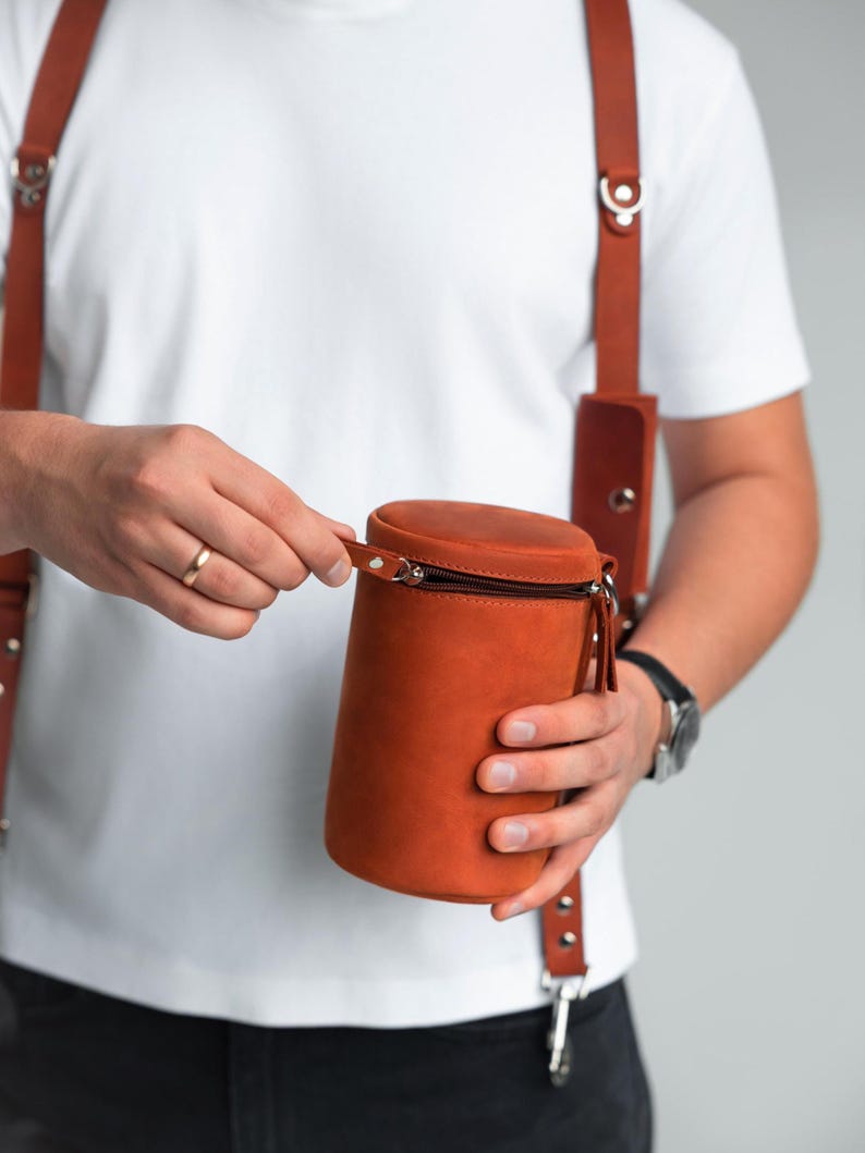 May include: A cylindrical, brown leather bag with a black zip is held by a person. The bag is worn with brown leather braces. The person is wearing a white t-shirt and a watch with a black strap.