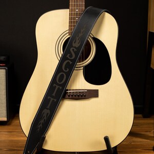 May include: Acoustic guitar with a black leather strap. The strap is embroidered with the name "SCOTT" in large letters and flame designs. The guitar has a light wood finish and a black sound hole.