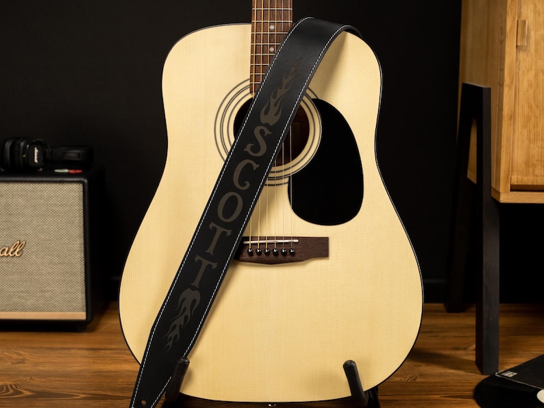 May include: Acoustic guitar with a black leather strap. The strap is embroidered with the name "SCOTT" in large letters and flame designs. The guitar has a light wood finish and a black sound hole.