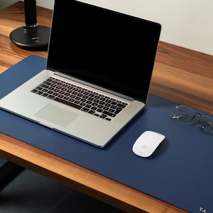 Op de afbeelding: Een marineblauwe bureaumat met een laptop, muis en bril op een houten bureau. De laptop is zilver en zwart, en de muis is wit. De bril heeft een dun, zilveren montuur. De bureaumat heeft de initialen "T.A.".