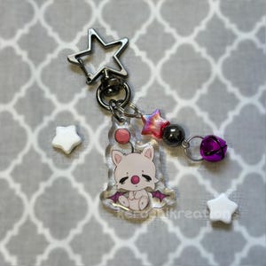 May include: A clear acrylic keychain featuring a white cartoon character with purple bat wings, a pink nose, and a red sphere. The keychain has a star-shaped clasp, a pink and red star bead, a dark gray bead, and a purple bell. The text "retro inkreations" is visible.