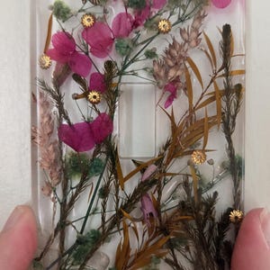 May include: A clear resin light switch cover with embedded dried flowers and a dragonfly charm. The flowers are various shades of pink, green, and brown, with gold accents. The switch opening is rectangular.
